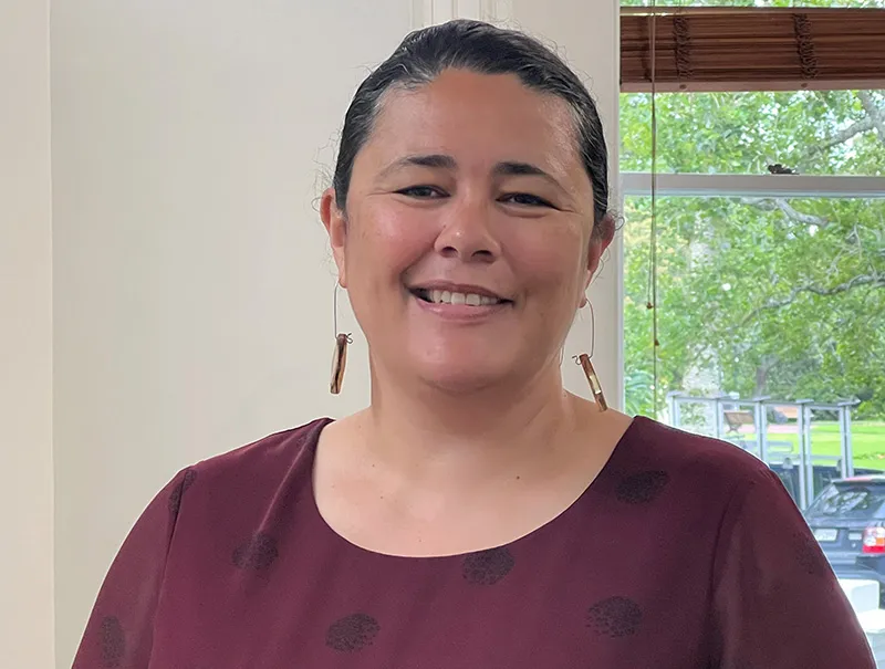 A smiling Dr Sandra Hanchard wears a maroon coloured blouse and dangly earrings. She stands in front of a window that looks out onto a tree and lawn. She is in a hospital environment.