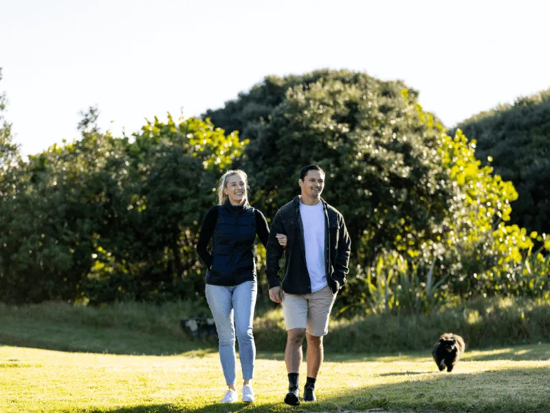 A couple walks arm-in-arm through a sunny, grassy park, smiling and enjoying the outdoors. The woman wears a black vest and jeans, while the man wears a jacket and shorts. In the background, lush green trees frame the scene, and a small black dog follows