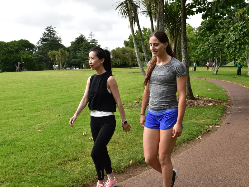 Two women walking on a park path surrounded by green grass and trees, exercising outdoors on a cloudy day