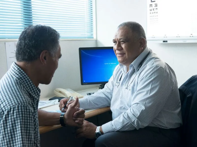 A doctor sits at a desk in a medical office, smiling and warmly holding the hand of a male patient during a consultation. The doctor wears a stethoscope around his neck, and a computer monitor, eye chart, and medical equipment are visible in the backgroun
