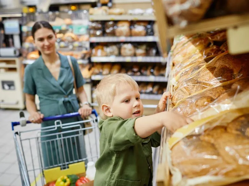 Young boy reaching for packaged bread on a supermarket shelf with his mother shopping in the background.