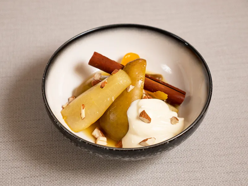 Image shows a bowl filled with poached pear halves, a cinnamon stick and a dollop of yoghurt. There are scattered almond slivers on top.