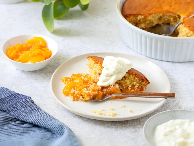 Apricot pudding with apple and almond sponge served warm on a plate with whipped cream and extra stewed apricots in bowls in the background.