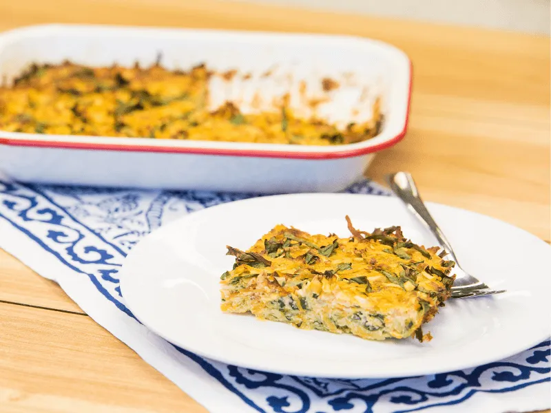 Golden baked vegetable frittata slice on a white plate with a fork, with the remaining casserole in a baking dish behind it.