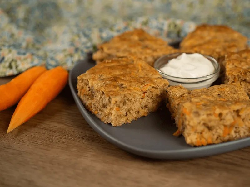 Homemade carrot and banana savory slices served on a gray plate with a small bowl of white dipping sauce in the center, accompanied by fresh carrot pieces on a wooden surface with a colorful fabric in the background.