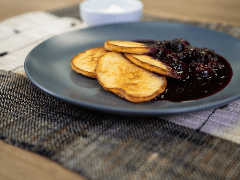 Banana pikelets served with a glossy mixed berry compote on a dark plate, with a small bowl of yogurt in the background.