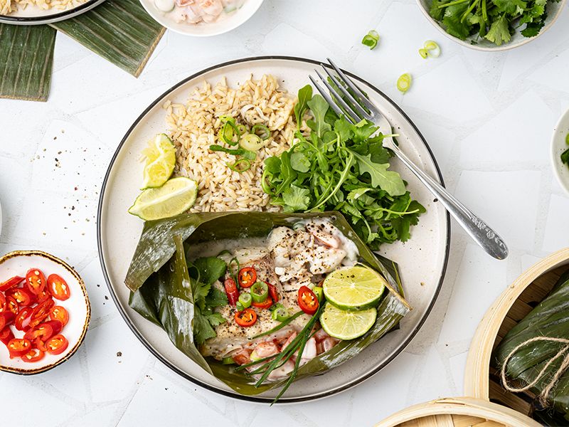 Steamed white fish fillet wrapped in banana leaf, garnished with sliced red chili, lime wedges, fresh herbs, served with brown rice and arugula salad on a plate.