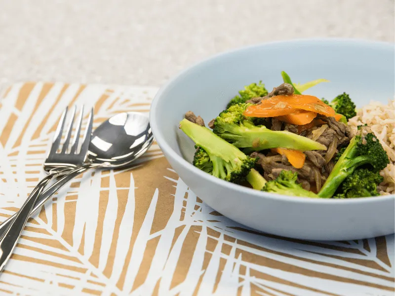 Beef and broccoli stir‑fry with carrot strips served with brown rice in a light blue bowl, set beside a fork and spoon.