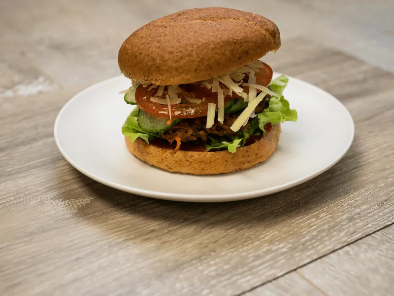 Classic beef burger with a lettuce‑topped patty, sliced tomato and cucumber, shredded cheese, and ketchup in a wholemeal bun on a white plate.