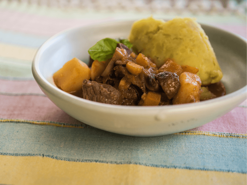 Hearty beef and vegetable stew with carrots, mushrooms, and potatoes, served alongside a scoop of creamy mashed potatoes in a shallow bowl.