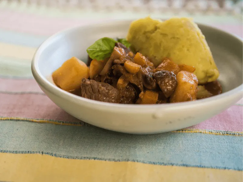 Hearty beef and vegetable stew with carrots, mushrooms, and potatoes, served alongside a scoop of creamy mashed potatoes in a shallow bowl.