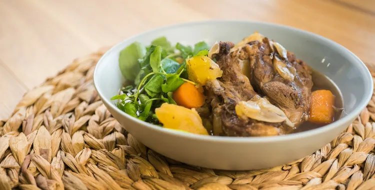 Traditional Māori boil‑up with pork bones, potatoes, carrots, and watercress in a light broth, served in a shallow bowl on a woven placemat.