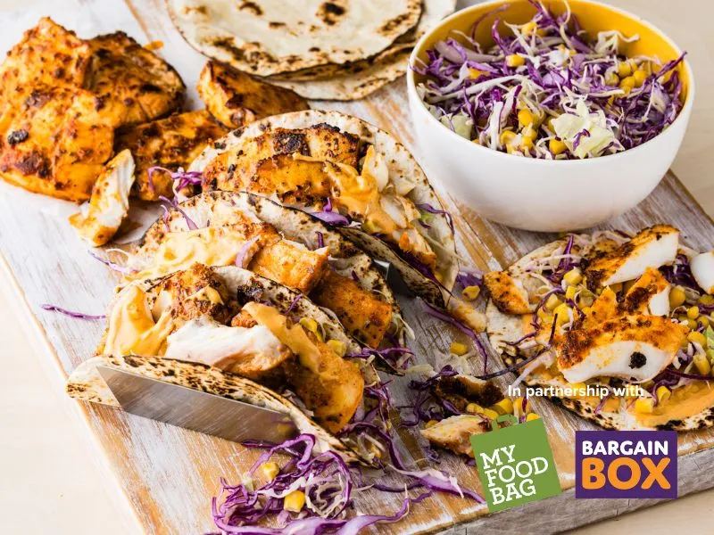 Alt text: Grilled Cajun fish tacos filled with sliced spicy fish, creamy orange sauce, and shredded purple cabbage on toasted tortillas, served on a wooden board with a bowl of corn and cabbage slaw and My Food Bag and Bargain Box logos in the corner.