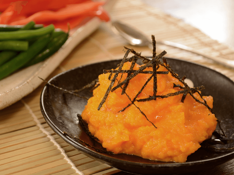 Bright orange carrot and miso dip topped with shredded nori on a black plate, with green beans and red peppers in the background.
