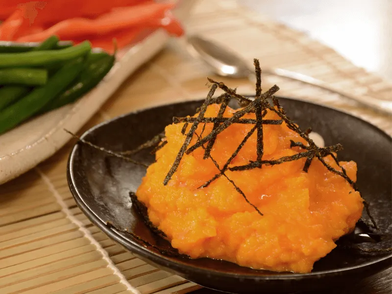 Bright orange carrot and miso dip topped with shredded nori on a black plate, with green beans and red peppers in the background.