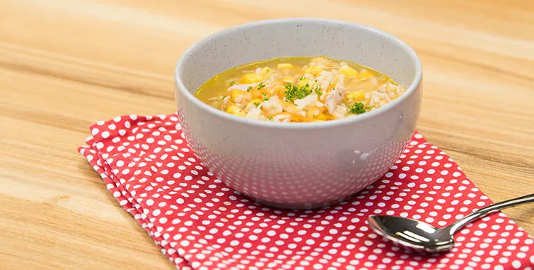 Carrot, chicken, and sweet corn soup in a white bowl, garnished with parsley, on a red polka dot napkin with a spoon beside it.