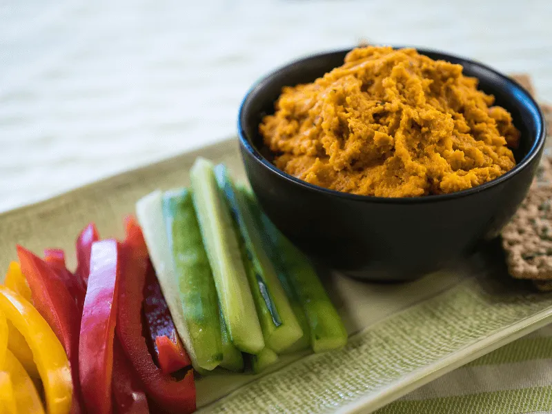 Carrot and cumin spread served in a black bowl with cucumber sticks, red and yellow capsicum, and crispbread on a rectangular platter.