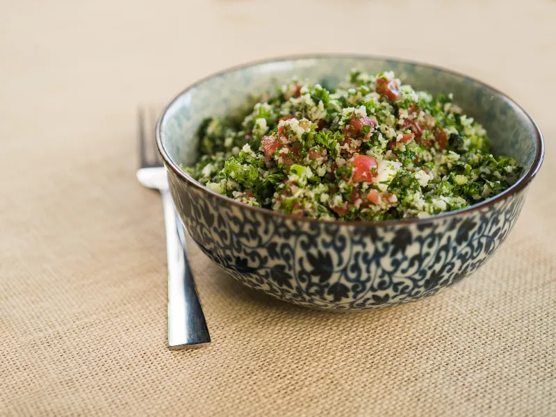 Bowl of cauliflower tabbouleh with parsley and tomato on a beige tablecloth.