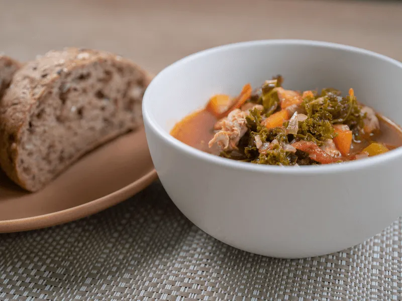 Chicken and kale soup with carrots in a tomato‑tinged broth, served alongside slices of whole‑grain bread in a white bowl.