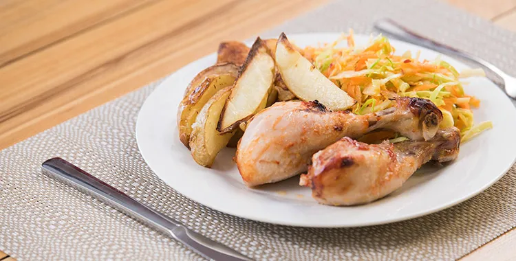 Roasted chicken drumsticks with potato wedges and tangy coleslaw on a white plate, set on a neutral placemat with cutlery.