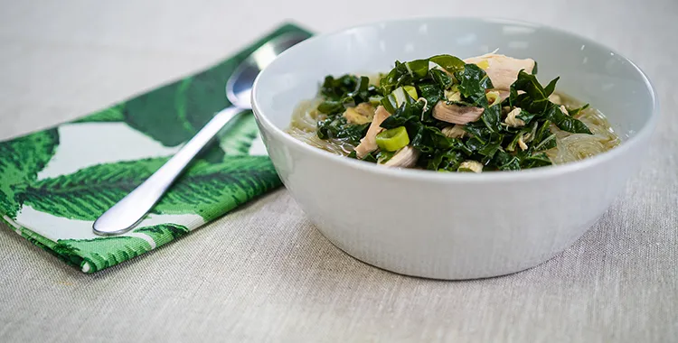 Chicken noodle soup with shredded chicken, greens, and glass noodles in a light broth, served in a white bowl with a spoon and napkin.