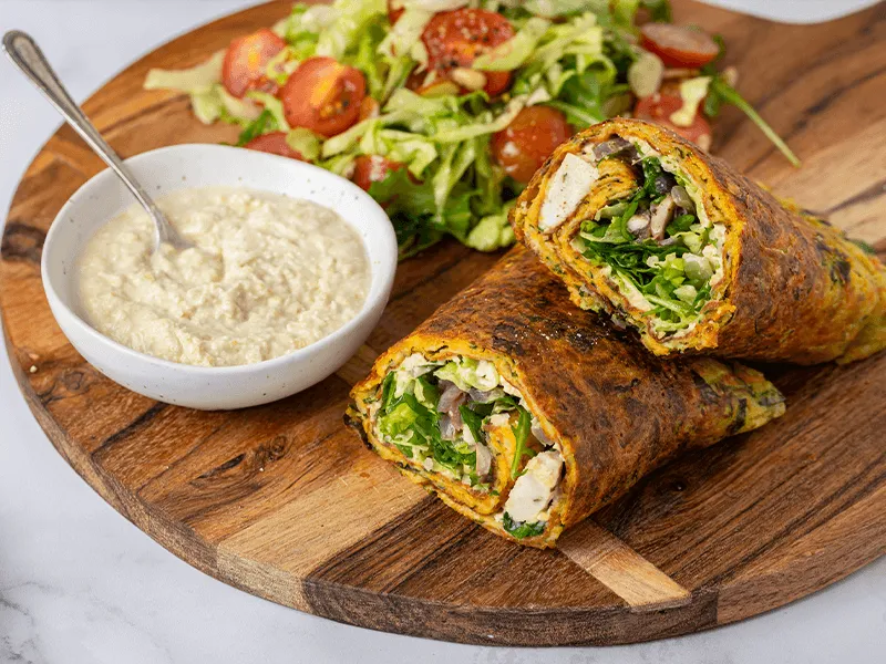 Chicken wrap (outer wrap made from vegetables) filled with sliced cooked meat, tofu, beans and cheese, served on a wooden platter with creamy dip and a side salad of lettuce and cherry tomatoes.