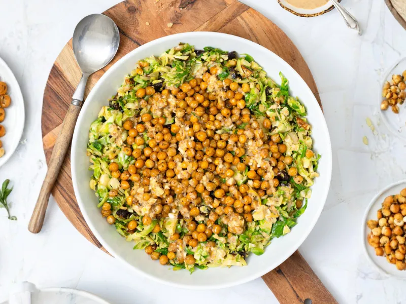 Overhead view of shaved Brussels sprouts salad topped with crispy roasted chickpeas and creamy lemon tahini dressing in a white bowl on a wooden board.