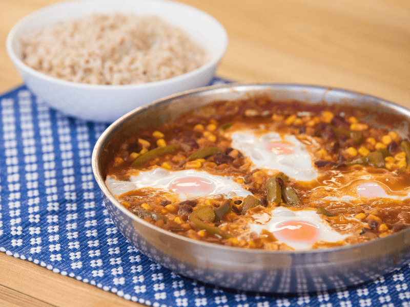 Skillet of chili beans with corn and peppers topped with baked eggs, served alongside a bowl of brown rice on a blue patterned mat.