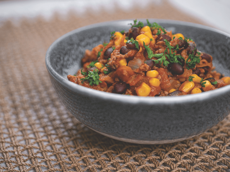 Chilli con carne with minced beef, black beans, sweetcorn, onions, and tomatoes, simmered with chili spices and garnished with parsley in a gray bowl.