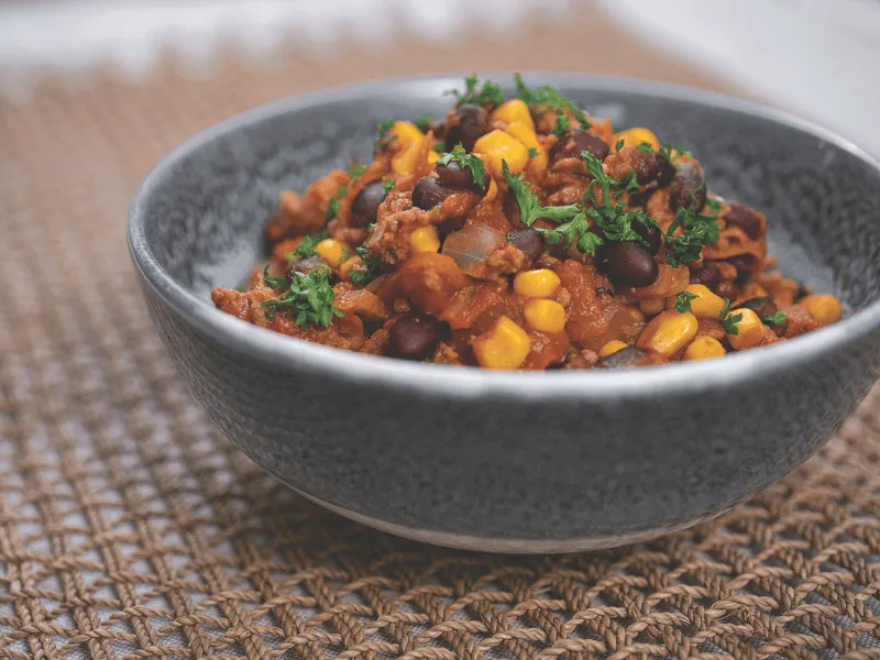 Chilli mince with beans and corn—ground beef chilli con carne simmered with tomatoes, black beans, sweetcorn, onions, and spices, garnished with parsley in a gray bowl.