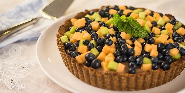 Fruit tart topped with blueberries and small cubes of melon and green fruit, garnished with a mint sprig on a white plate