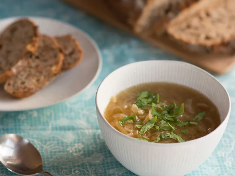 Rustic classic onion soup in a white bowl, topped with fresh herbs, served with slices of crusty bread on the side.