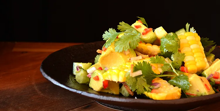 Fresh corn and avocado salad with cilantro, red onion, and chili in a black bowl on a wooden table.
