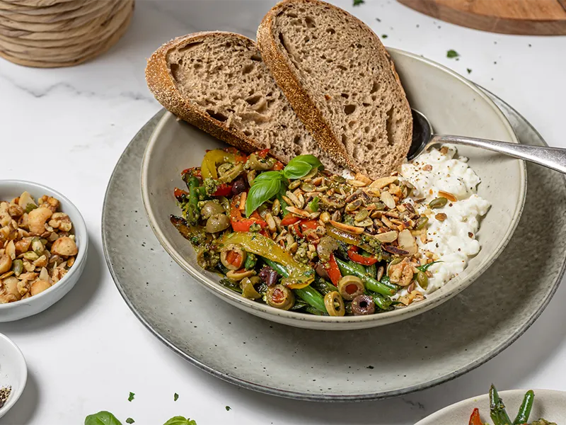 Image of a heart-healthy cottage cheese bowl topped with BBQ green beans, capsicum, olives, pickled onion, pesto and chopped nuts, served with toasted wholegrain bread.