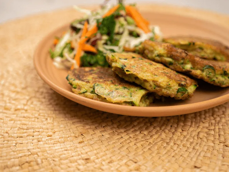 Golden courgette fritters—crispy zucchini patties with herbs—served with a fresh slaw salad on a tan plate.