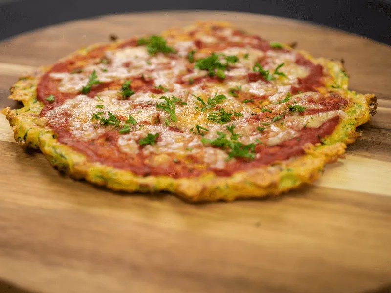 Courgette pizza with a grated‑zucchini crust topped with tomato sauce, melted cheese, and parsley, served on a wooden board.