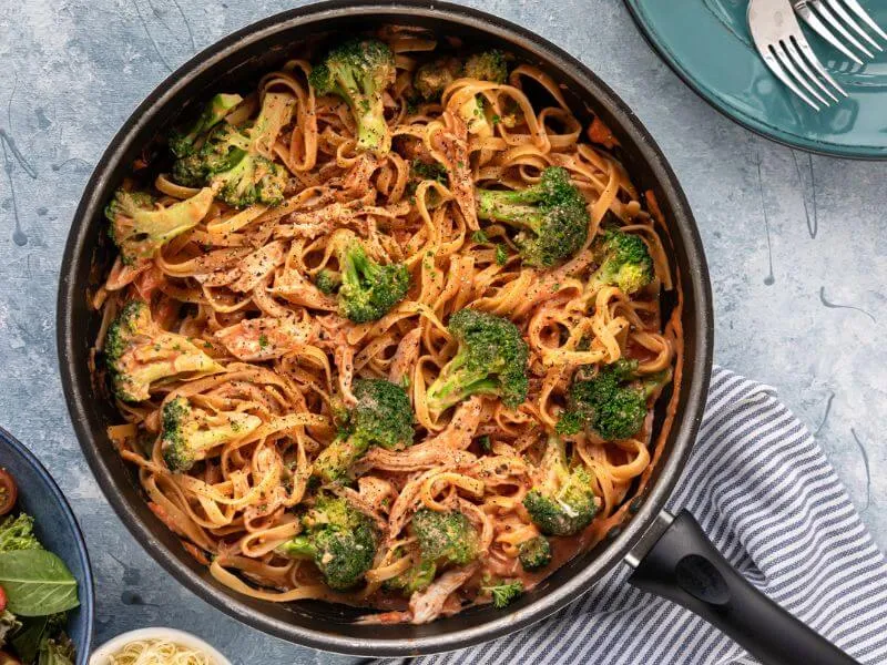 A brown bowl against a brown tablecloth holds a serving of rigatoni pasta (large pasta tubes) covered in an orange coloured sauce (creamy tomato).