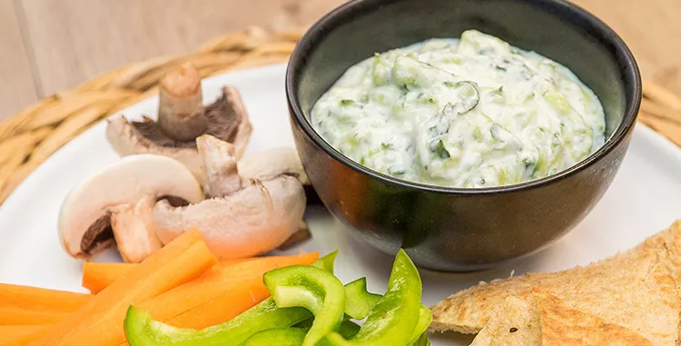 Cucumber and yoghurt dip (tzatziki) in a black bowl served with carrot sticks, green capsicum, mushrooms, and toasted pita on a platter.