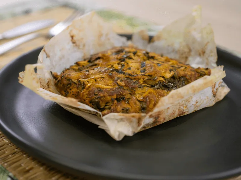 Curried pork lap‑lap baked in parchment, a Vanuatu‑inspired dish of grated root vegetables with pork and spices, set on a dark plate.