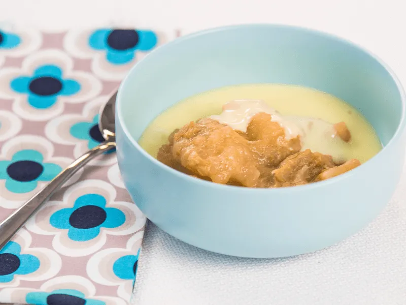 Warm custard with stewed fruit and a dollop of yogurt served in a light blue bowl beside a patterned napkin and spoon.
