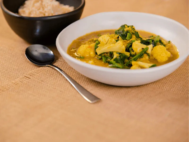 Yellow lentil dhal curry with cauliflower and spinach in a white bowl, served with a side of brown rice on a textured cloth.