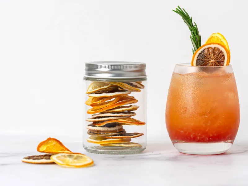 Glass jar of dried citrus slices next to a refreshing orange cocktail garnished with dehydrated citrus and rosemary on a white background.