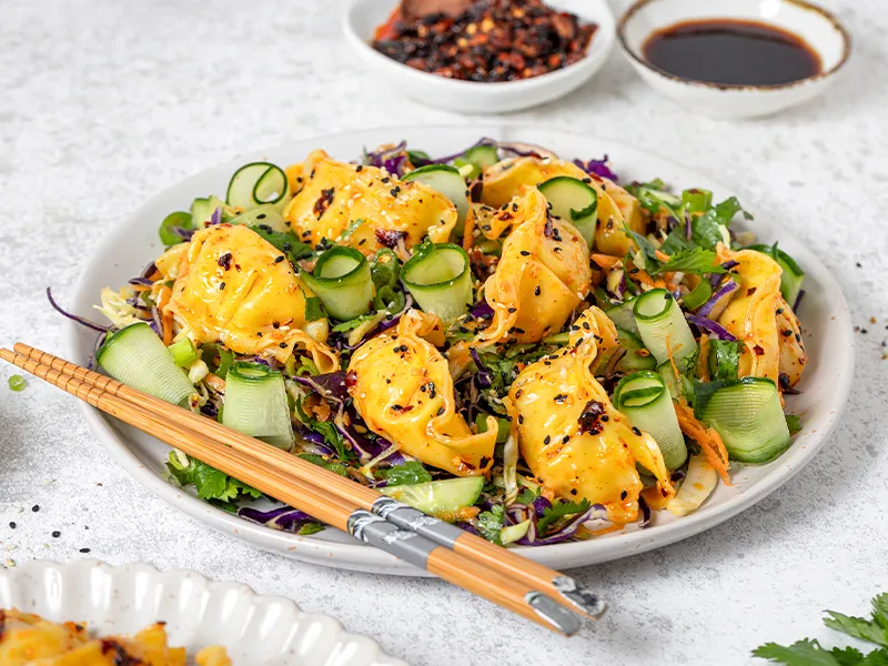 Asian dumplings on fresh salad with cucumber, carrots, purple cabbage, and sesame seeds, garnished with chili flakes, served with chopsticks and dipping sauce.