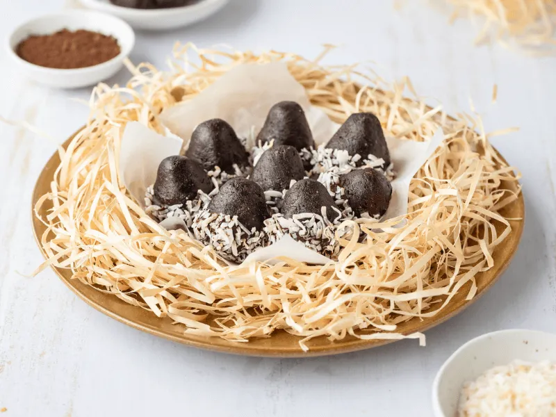 Chocolate coconut Easter eggs arranged on parchment paper in a nest of shredded straw, with some eggs coated in desiccated coconut, served on a wooden plate.