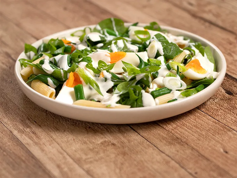 A white plate on a wooden surface holds a serving of egg and bean pasta salad.
