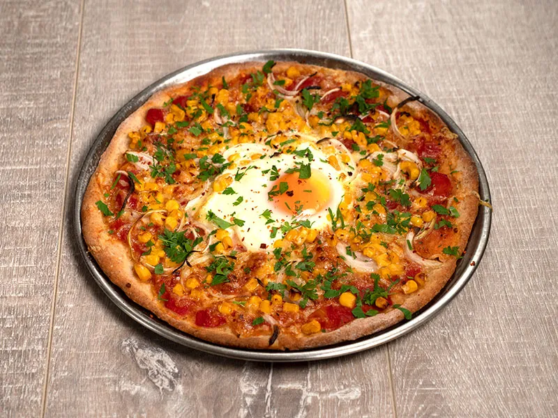 A whole spicy egg and corn pizza is displayed in a metal tray on a medium brown, wooden surface.