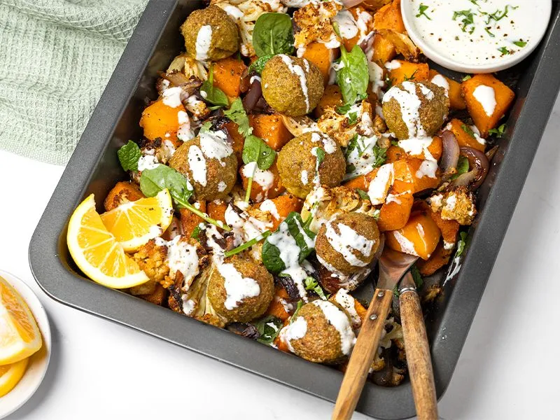 A top-down view of a rectangular baking tray filled with roasted falafel, cauliflower, fennel and pumpkin and drizzled with a lemony yoghurt tahini dressing.