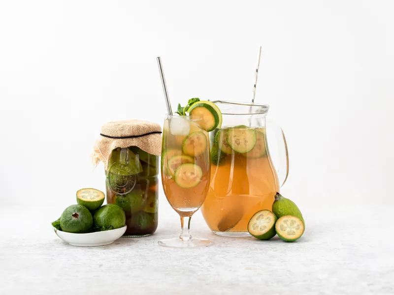 A glass and a pitcher of feijoa-infused drink with ice and sliced feijoas, next to a jar of preserved feijoas and a small bowl of whole and halved feijoas, all arranged on a light surface with a white background.
