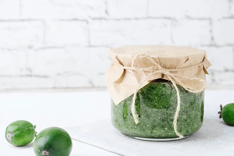 A jar of fresh, homemade feijoa salsa sits on a white kitchen bench in front of white brick wall. Scattered around the jar are whole feijoa fruits.