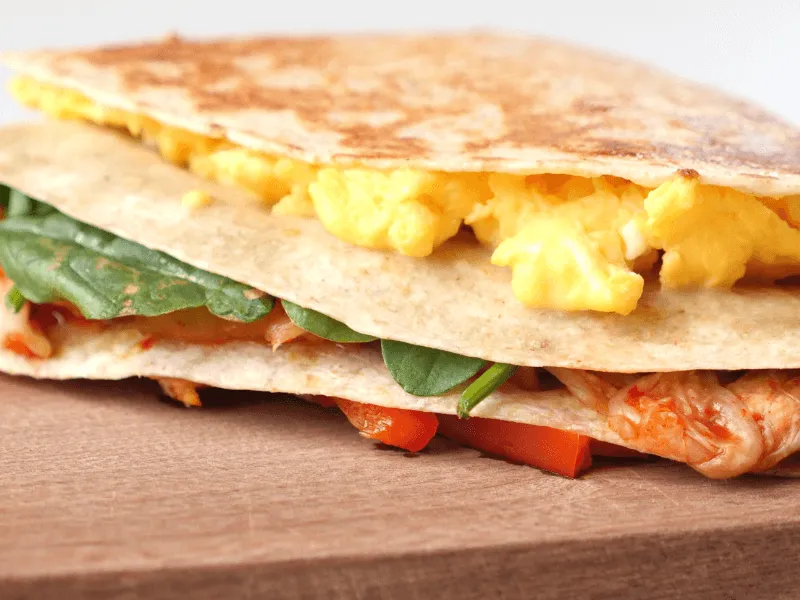 Breakfast quesadilla with scrambled eggs, spinach, chicken, and red bell peppers on a wooden board.